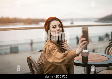 Schöne junge Frau nimmt Selfie mit ihrem Smartphone, während mit einer Tasse Kaffee. Französin trägt rote Baskenmütze sitzt auf der Terrasse des Restaurants oder Stockfoto