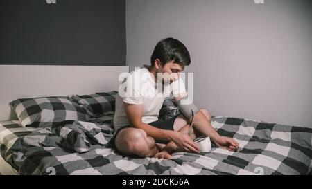 Junge kaukasische Mann misst den Blutdruck zu Hause im Schlafzimmer sitzen auf dem Bett zu Hause Kleidung. Männlich verwendet medizinische Instrument Tonometer, um seine eigenen zu überprüfen Stockfoto