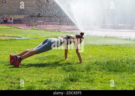 Junge Frau macht einen Liegestütze und Stretching Fitness-Training im örtlichen öffentlichen Sportstadion, während ein automatischer Sprinkler das Gras bewässert. Lif Stockfoto