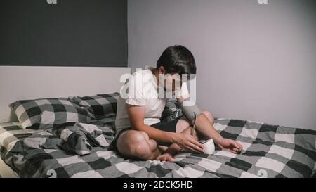 Junge kaukasische Mann misst den Blutdruck zu Hause im Schlafzimmer sitzen auf dem Bett zu Hause Kleidung. Männlich verwendet medizinische Instrument Tonometer, um seine eigenen zu überprüfen Stockfoto