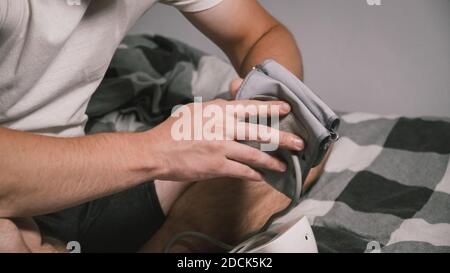 Junge kaukasische Mann misst den Blutdruck zu Hause im Schlafzimmer sitzen auf dem Bett zu Hause Kleidung. Männlich verwendet medizinische Instrument Tonometer, um seine eigenen zu überprüfen Stockfoto