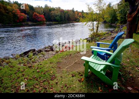 Zwei Adirondack-Stühle, die an einem Flussufer mit Bäumen sitzen Im Hintergrund Stockfoto