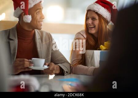 Junge afro-amerikanische Mitarbeiterin trinkt Kaffee im Büro im Gespräch mit kaukasischen Kollegin, trägt santa Hüte. weihnachtszeit Stockfoto