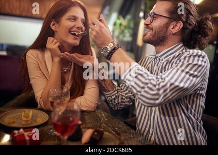 Ein junger Mann füttert seine Freundin am Valentinstag Feier in einem Restaurant in einer fröhlichen Atmosphäre. Gemeinsam, Jubiläum, Feier Stockfoto