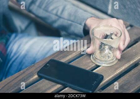 Nahaufnahme von leerem Kaffeeglas und schwarzem Smartphone auf Holztisch der Cafeterrasse. Suchtkonzept, Freizeit Stockfoto