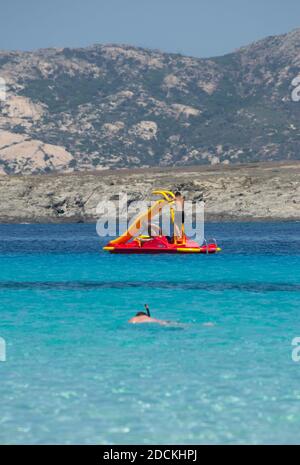 Zwei Personen genießen ein Tretboot, während ein junger Mann Schnorcheln im türkisfarbenen und klaren Meer von ​​sardinia Stockfoto