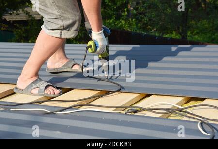 Ein Bauunternehmer installiert Kastenprofilbleche, lackierte Metalldachbleche auf dem Dach des Hauses mit einem elektrischen Schraubendreher. Stockfoto