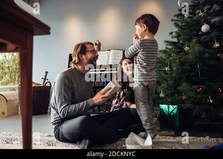 Eltern schenken ihrem Sohn, der neben einem Weihnachtsbaum sitzt. Junge sieht aufgeregt, während immer Weihnachtsgeschenk. Stockfoto