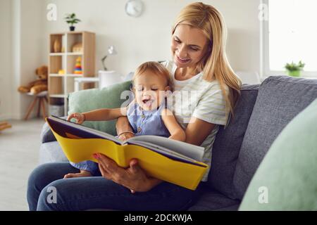 Glückliche Mutter mit lächelndem Baby auf den Knien und Buch lesen Zu Hause