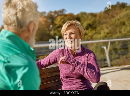 Fröhliches, sportliches Seniorenpaar, das sich nach dem Training im Freien auf der Bank ausruhte. Reife Frau lächelt zum Mann, während sie etwas diskutiert Stockfoto