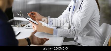 Unbekannte junge Ärztin hört ihrer Patientin zu, während sie in der Klinik zusammen sitzt. Medizinkonzept während der Coronavirus-Pandemie Stockfoto