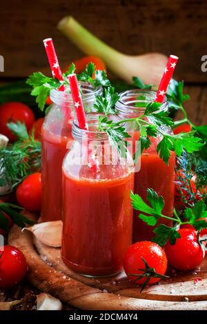 Würziger Tomatensaft in kleinen Flaschen mit Strohhalmen, Kirschtomaten, Petersilie, Dill, Knoblauch, Gewürzen und Salz, selektiver Fokus Stockfoto