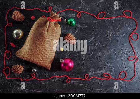 Weihnachtstasche aus grobem Sackleinen, Zapfen, Weihnachtsschmuck, Rahmen aus rotem Garn auf dunklem Hintergrund. Speicherplatz kopieren. Stockfoto