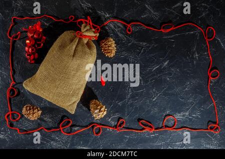 Weihnachtstasche aus grobem Sackleinen, Zapfen, Weihnachtsschmuck, Rahmen aus rotem Garn auf dunklem Hintergrund, Platz für Text. Speicherplatz kopieren. Stockfoto