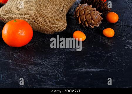 Weihnachtstasche aus grobem Sackleinen, Zapfen, Mandarinen oder Orangen, Kumquat auf dunklem Hintergrund. Speicherplatz kopieren. Stockfoto