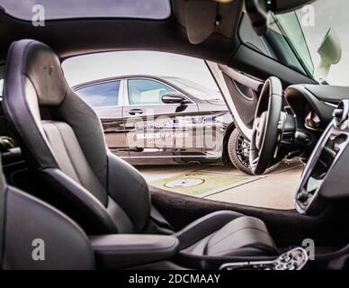 Innendetail eines Luxus-Leistungsfahrzeugs mit cremeweißen Ledersitzen. Durch das Fahrzeugfenster zeigt die Reflexion einen schwarzen Brabus VIP Shuttle Mercedes, der an einer Flughafeneinrichtung geparkt ist. Professionelle Automobilfotografie, die die Handwerkskunst im Innenraum und die Präsenz der Flotte im Freien betont. Stockfoto