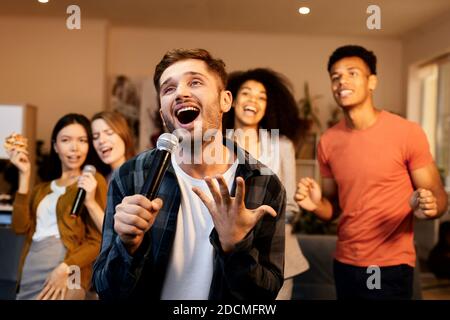 Spaß zu Hause. Enthusiastisch schöner junger Mann, der mit Mikrofon singt, während er mit Freunden im Haus Karaoke spielt und in der modernen Wohnung steht Stockfoto