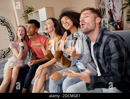 Genießen Sie einfach den Moment. Gruppe von multikulturellen Freunden, die enthusiastisch beim Karaoke spielen zu Hause, mit Mikrofon singen, auf dem sitzen Stockfoto