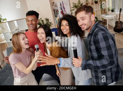 Machen wir Karaoke. Gruppe von multikulturellen Freunden, die begeistert, singen mit Mikrofon, während Karaoke zu Hause spielen, in der modernen stehen Stockfoto