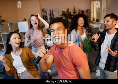 Genießen Sie Spaß. Enthusiastischer junger Mann, der mit Mikrofon singt, während er Karaoke mit Freunden in der modernen Wohnung spielt und eine Heimparty hat Stockfoto