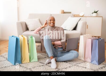 Glückliche schwarze Dame in Hijab sitzt auf dem Boden mit Laptop und Papiertaschen, Einkaufen im Internet zu Hause Stockfoto