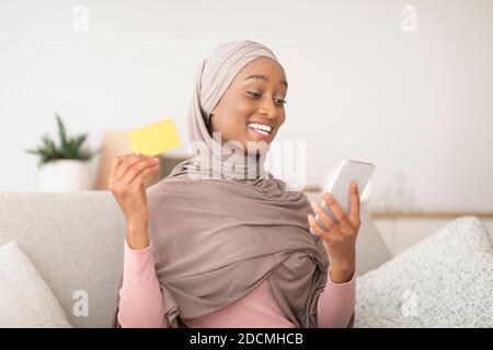 Einkaufen zu Hause. Schwarze Dame in Hijab hält Kreditkarte und Smartphone, so dass Online-Kauf auf Sofa drinnen Stockfoto