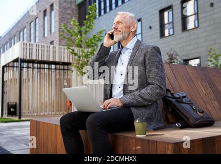 Aufbau starker Beziehungen. Beschäftigt elegante reifen Mann am Telefon sprechen, arbeiten auf seinem Laptop, während auf der Bank im Freien sitzen Stockfoto