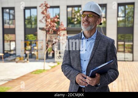 Städtisches Design. Erfolgreicher Architekt mittleren Alters, Ingenieur Manager trägt Helm lächelnd, im Freien stehend und hält eine Blaupause in seinem Stockfoto