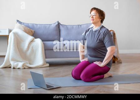 Ältere Frau meditiert. Sie sitzt auf den Knien vor dem Monitor. Gesundes Lifestyle-Konzept. Stockfoto