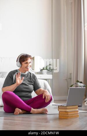 Ältere Frau mit Kopfhörern bereitet sich auf das Training im Internet vor. Sie grüßt ihren Trainer. Stockfoto