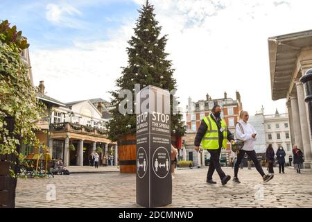 Die Menschen gehen an einem Stop vorbei das Schild mit der Verbreitung des Coronavirus neben dem Weihnachtsbaum in Covent Garden, London. England wird ein hartes Tier-System durchsetzen, sobald die Sperre am 2. Dezember endet. Stockfoto