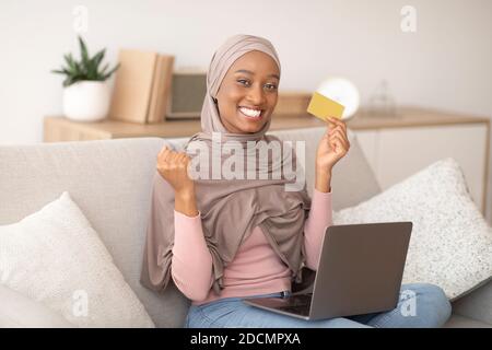 Glückliche schwarze Frau in Hijab hält Kreditkarte, so dass JA Geste, Online-Shopping auf Laptop-Computer zu Hause Stockfoto