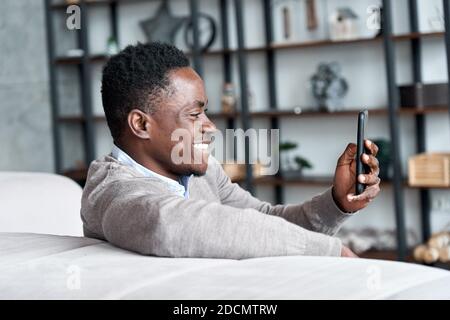 Glücklicher afrikanischer junger Mann, der mobile Apps verwendet, die zu Hause auf der Couch sitzen. Stockfoto