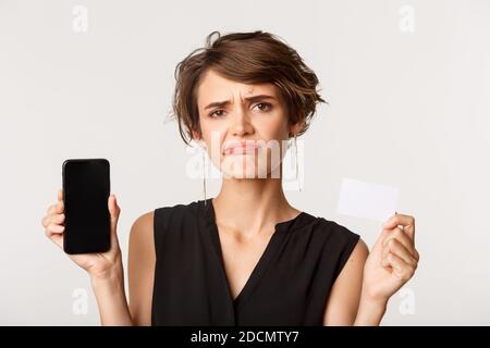 Nahaufnahme der enttäuschten, skeptischen Brünette Frau grinsen unamüsiert, zeigt Kreditkarte und Smartphone-Bildschirm, stehen auf weißem Hintergrund Stockfoto
