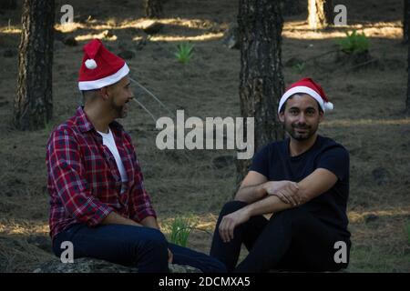 Junge Homosexuell Paar mit roten Weihnachtsmützen sitzen auf Kiefernwald Park. Zwei Männer posieren in der Winterferienzeit im Freien in der Natur Stockfoto