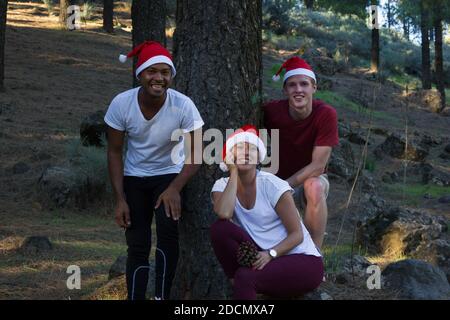 Drei lächelnde Freunde posieren mit roten Weihnachtsmützen im Kiefernwald Park. Multi ethnische junge Gruppe in T-Shirts glücklich für die Winterferienzeit Stockfoto