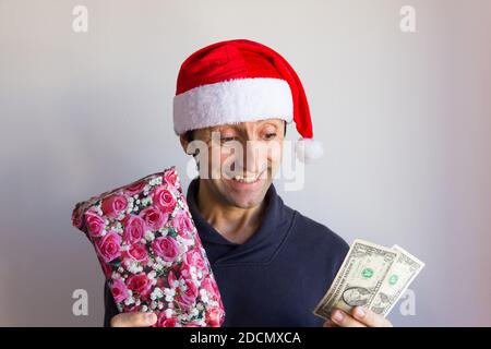 Lächelnder Mann mit Weihnachtsmann roten Hut hält eingewickelte Geschenk und Dollar Scheine. Sparen Sie Geld auf Weihnachtsgeschenke, kleines Budget für Winterurlaub Saison einkaufen Stockfoto