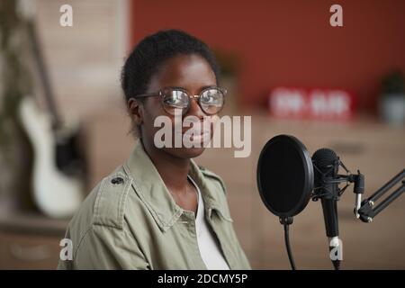 Porträt einer jungen afroamerikanischen Frau, die beim Sitzen am Mikrofon im Aufnahmestudio zu Hause auf die Kamera schaut, Raum kopieren Stockfoto