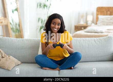 Spaß bleiben zu Hause Aktivitäten. Lächelnde schwarze Frau mit Joystick spielen Online-Videospiele auf Couch, voller Länge Porträt Stockfoto