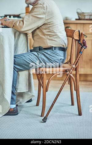 Zugeschnittenes Foto eines älteren Mannes, der am Tisch in der Küche mit Gehstock in seiner Nähe sitzt. Pflege- und Gesundheitskonzept Stockfoto