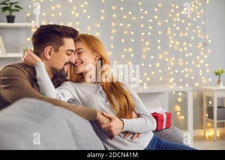 Dankbare Frau sitzt auf der Couch neben einem Weihnachtsgeschenk und umarmt und küsst ihren Mann. Stockfoto