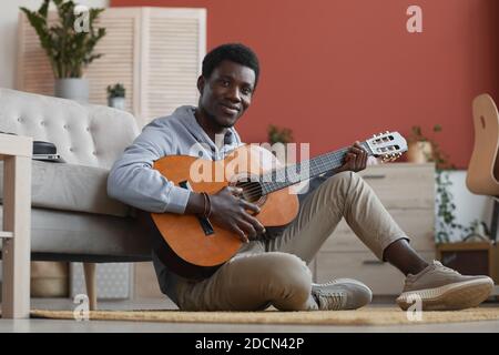 Porträt eines jungen afroamerikanischen Mannes, der Gitarre spielt und die Kamera anlächelt, während er zu Hause auf dem Boden sitzt und Platz kopiert Stockfoto