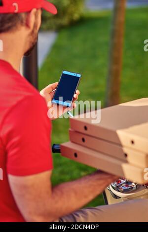 Rückansicht eines Liefermanns in roter Uniform, der auf einem Roller sitzt und mit dem Smartphone unterwegs ist, während er Pizza liefert. Konzept für die Lebensmittelzustellung Stockfoto