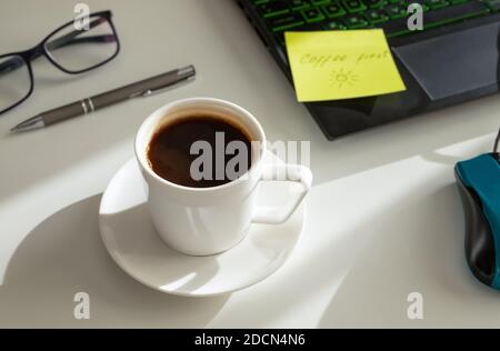 Eine Tasse Kaffee am Arbeitsplatz, ein Stift, ein Laptop, eine Brille. Kaffee zuerst Konzept. Hartes Licht. Selektiver Fokus. Horizontale Ausrichtung. Stockfoto