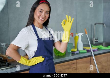 Portrait der fröhlichen jungen Frau, Putzfrau mit Schutzhandschuhen, lächelnd und zeigt OK-Zeichen, während sie sich bereit für die Reinigung des Hauses. Hausarbeit und Housekeeping, Reinigungsdienstkonzept Stockfoto