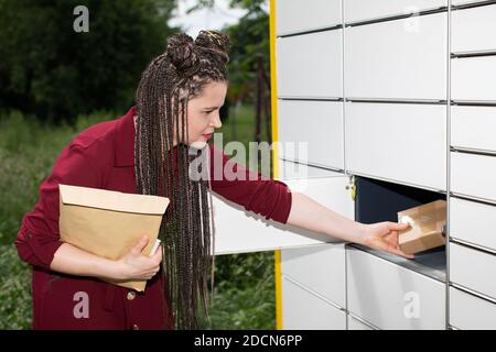 Ein junges Mädchen nimmt sorgfältig ein Paket für Paket aus dem Briefkasten. Automatischer Paketschrank in Lublin. Afrikanische Zöpfe. Stockfoto