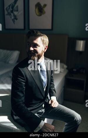 Ein flacher Fokus eines fröhlichen erwachsenen Mannes mit einem Bart trägt einen eleganten Anzug und sitzt auf einem Bett Stockfoto