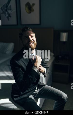 Ein flacher Fokus eines fröhlichen erwachsenen Mannes mit einem Bart trägt einen eleganten Anzug und sitzt auf einem Bett Stockfoto