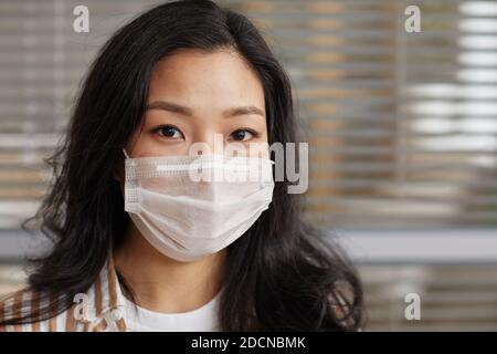 Nahaufnahme Porträt der jungen asiatischen Frau trägt Maske und Blick auf Kamera gegen Büro Jalousien Hintergrund, kopieren Raum Stockfoto