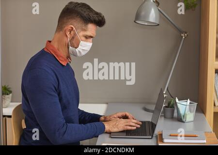 Seitenansicht Porträt von modernen reifen Mann trägt Maske und Mit einem Laptop am Schreibtisch im Büro arbeiten Stockfoto